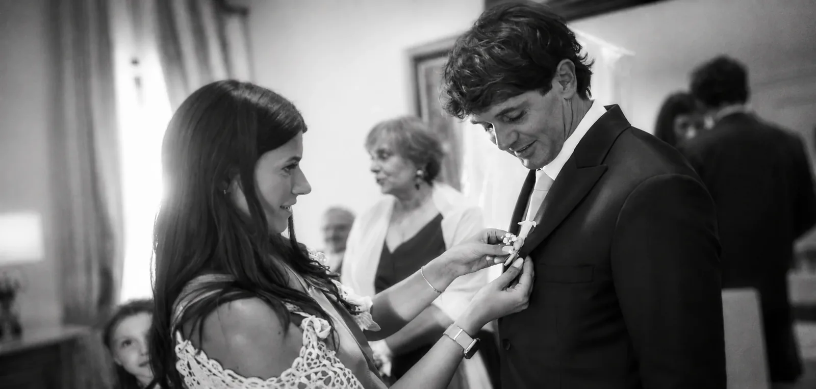 Una donna appunta il fiore all’occhiello allo sposo durante i preparativi del matrimonio, fotografia in bianco e nero