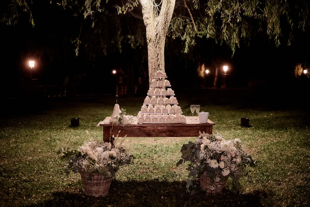 Torta nuziale illuminata di notte sotto un salice, con composizioni floreali ai lati